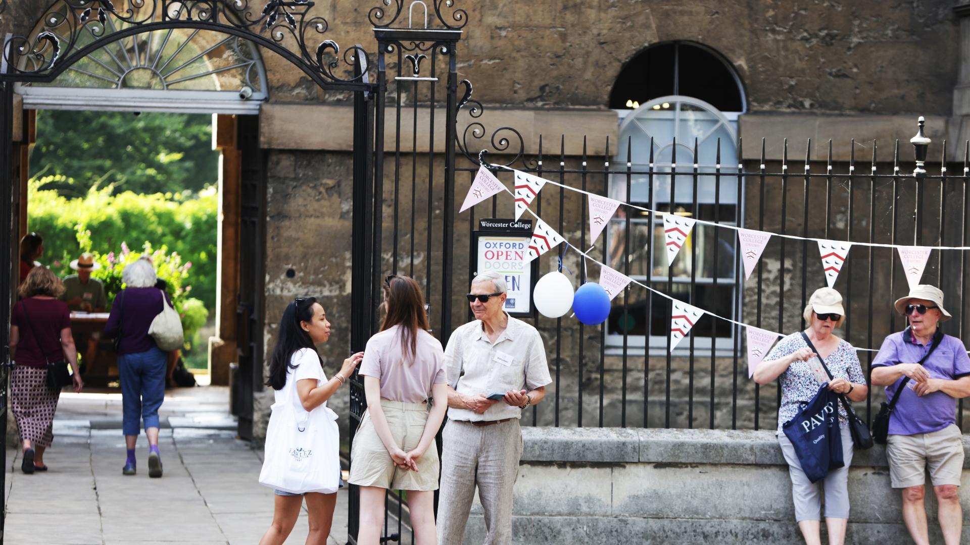 Two groups of people standing outside a historic stone building. An ironwork fence displays bunting.