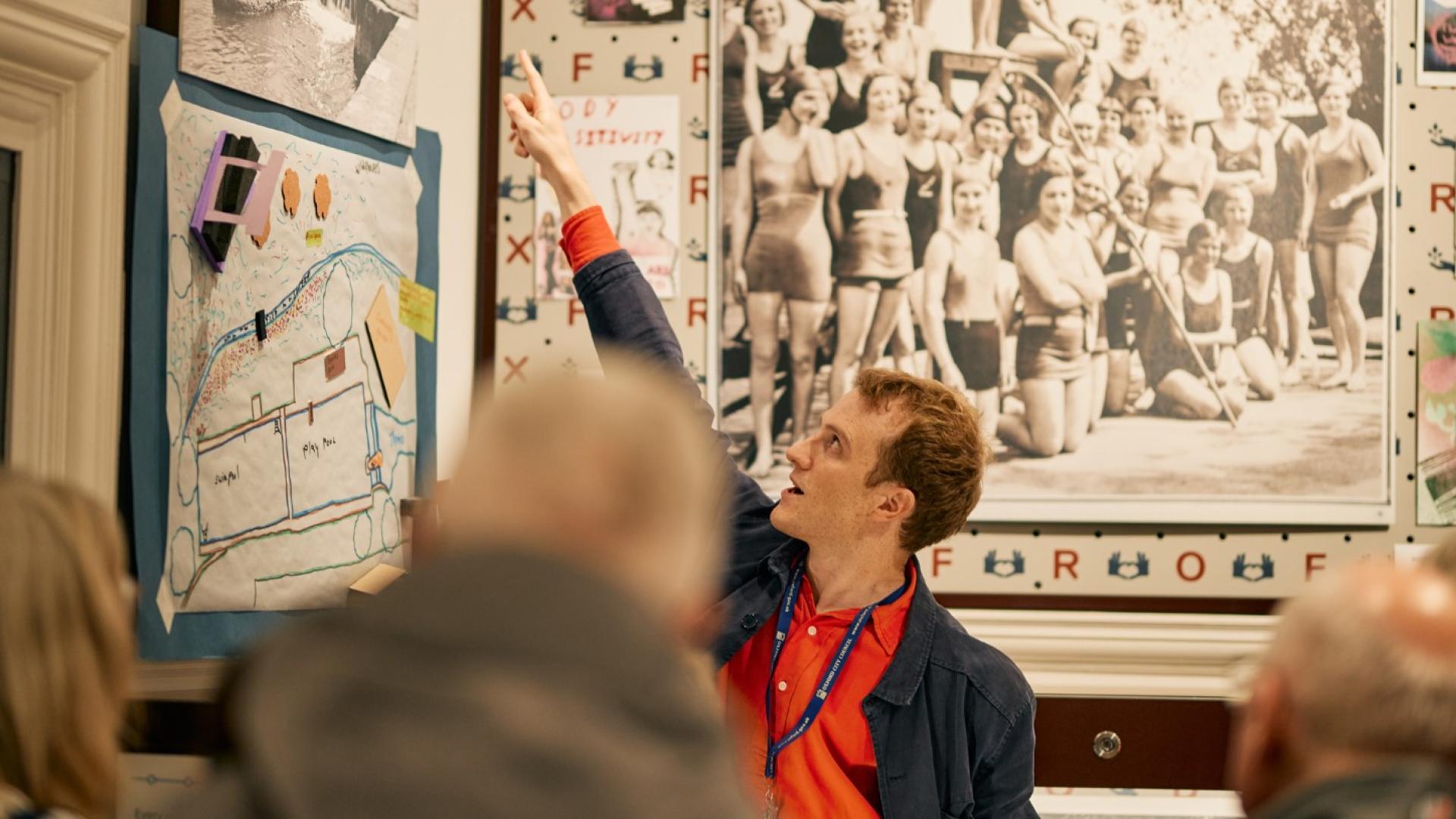 A guide in an orange shirt points at a history display during a tour to members of the public.