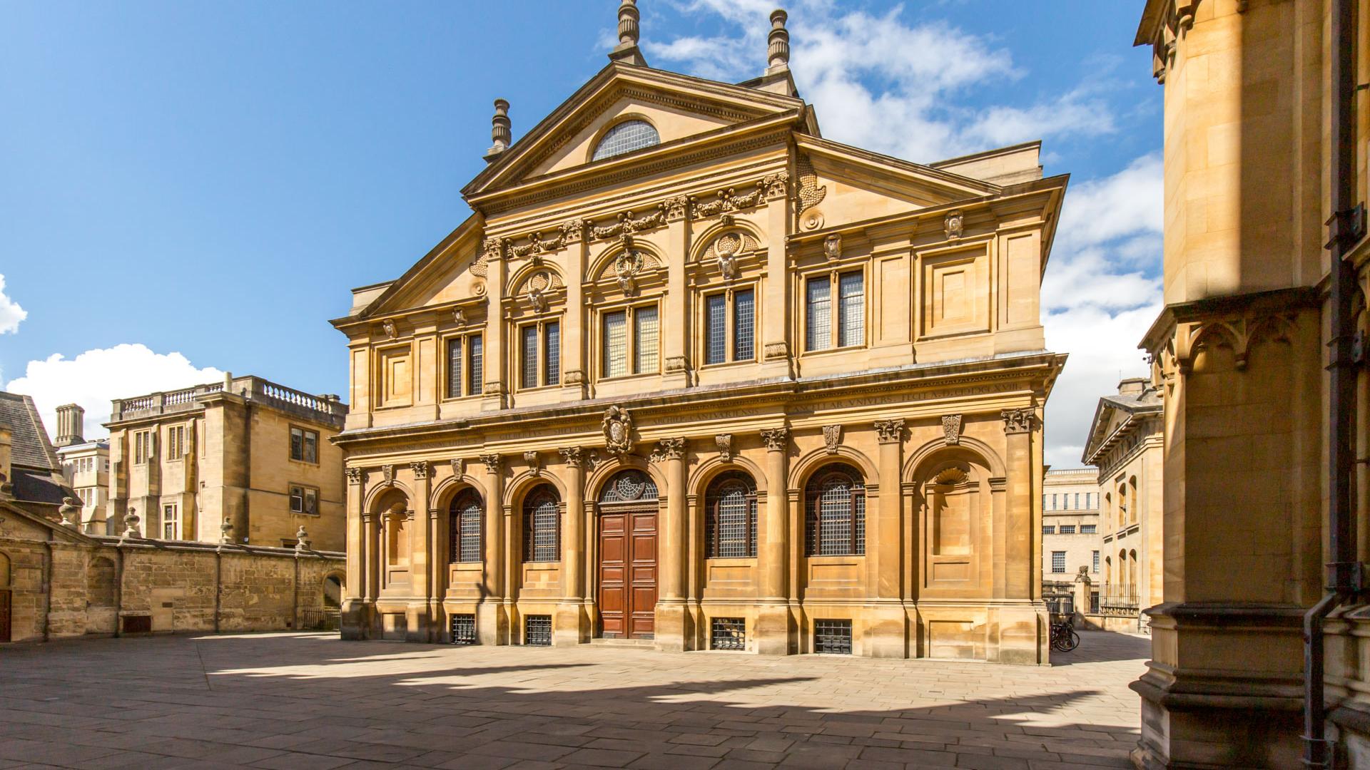The front of a sandy-coloured stone building with windows with rounded arches and a pediment.