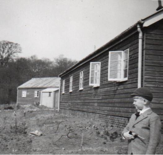 A black and white photograph of a child standing in front of wooden hut in a grassy area.