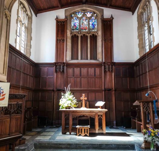 A wood-panelled church chancel with high windows on three sides and a wooden cross on a table in the centre.