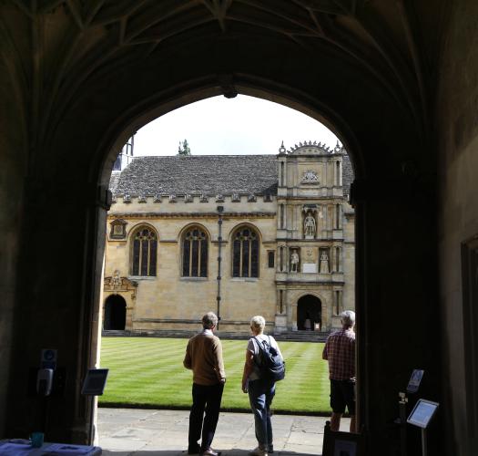 Picture of a dark strone archway of a gatehouse with two people looking out into a courtyard with another stone building in the background.