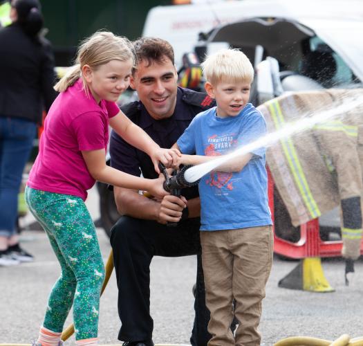 A firefighter crouching to help a boy and a girl direct a firehose.