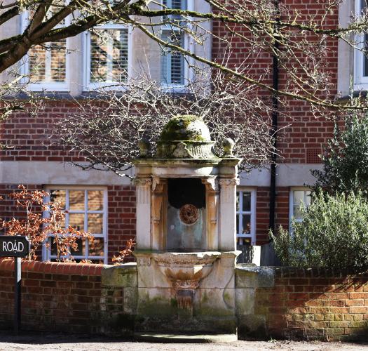 An ornate Victorian stone fountain set between two low brick walls, with trees overhead and brick houses in the background.