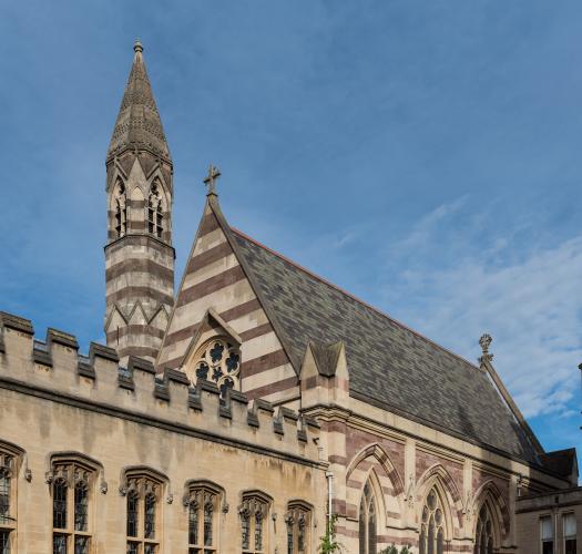 A building with a grey roof and walls striped with two different colours of stone. A tall stone tower points up into a blue sky.
