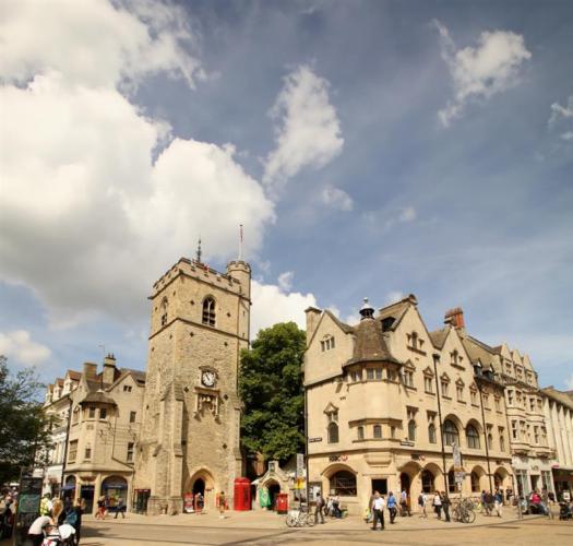 A church tower with a clock and crenellations at the top on a city street corner.