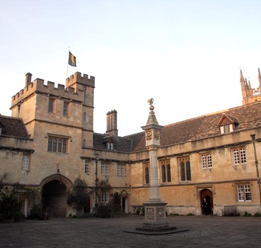 A sandy-coloured stone building with a gatehouse set around a paved courtyard with a column supporting a clock at the centre.