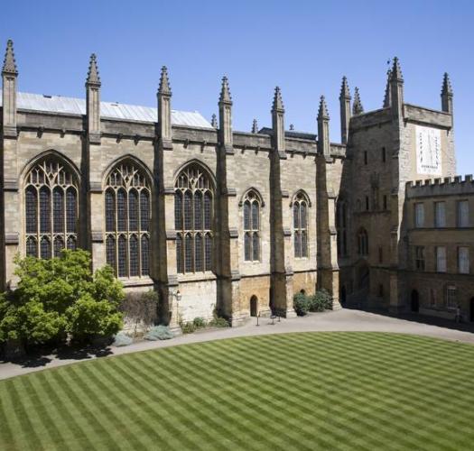 A stone chapel building with large windows and pinnacles, set around a oval lawn and against a bright blue sky.