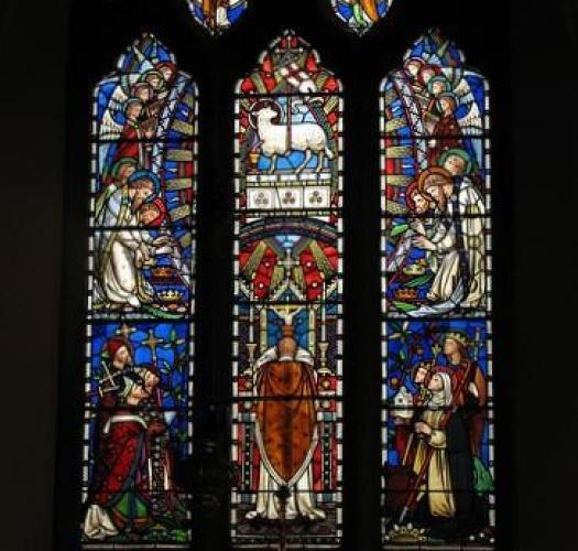 A stained glass window with a lamb, angels and saints, lit by sunlight in a dark church interior.