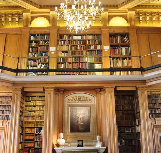 A library with tall bookcases, a balcony, an oil portrait of a man and a chandelier.