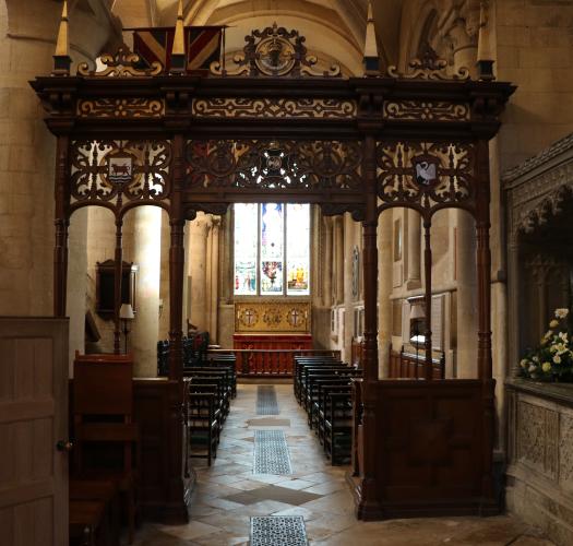 A carved wooden screen in a stone cathedral building with a clear arched window in the background. A stone tomb is visible to the right-hand side.