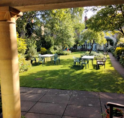 A garden with tables and chairs in dappled shade, viewed from beneath a stone portico.