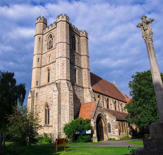 A stone church with a tower and terracotta-coloured roof tiles. A sculpted stone cross on a post is in the foreground.
