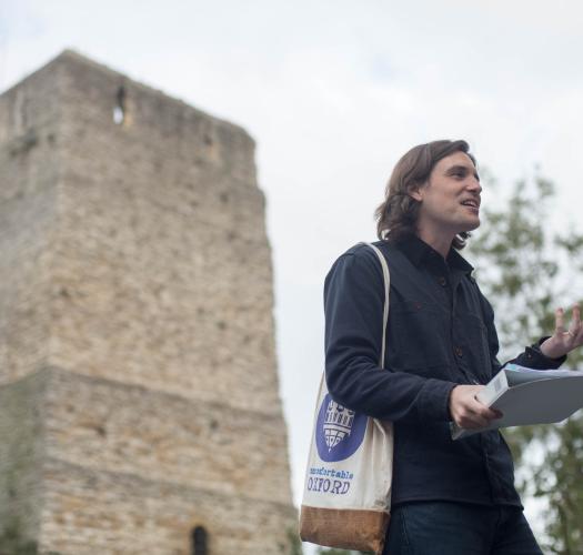 A tour guide with a tote bag that says Uncomfortable Oxford speaking in front of a square stone castle tower.