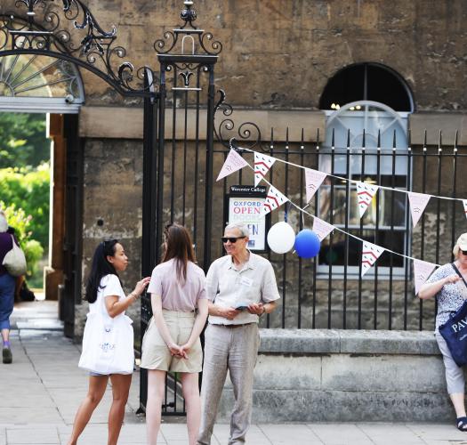Two groups of people standing outside a historic stone building. An ironwork fence displays bunting.