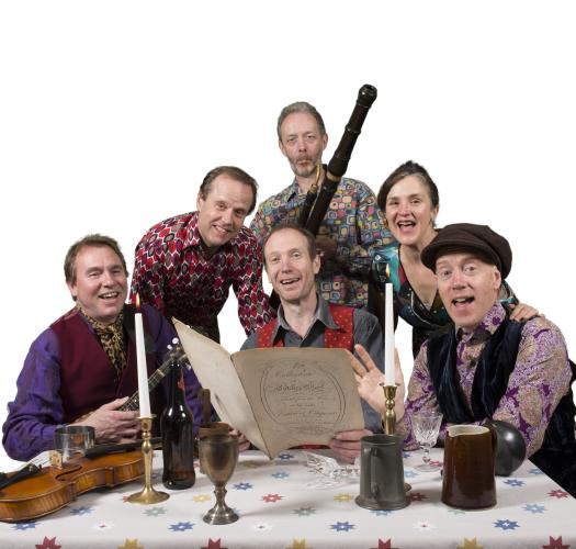 A group of musicians gather around a table with tankards, a candlestick and sheet music.