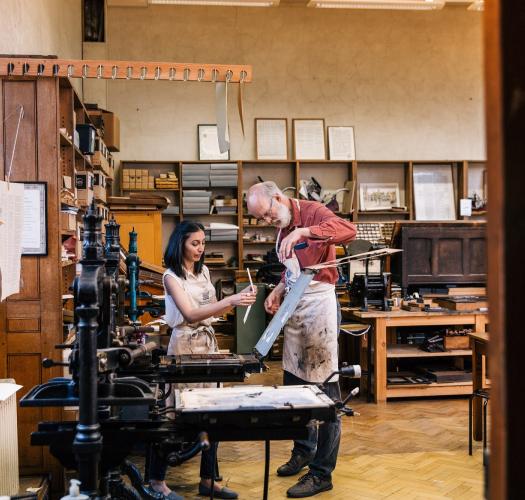 A man helps a woman operate a historic printing press in a workshop with wooden shelves.
