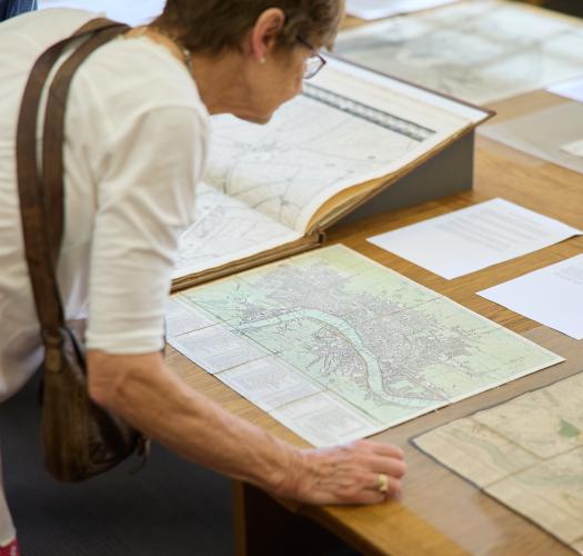 A women leans over a table to look at a series of historic maps on display.