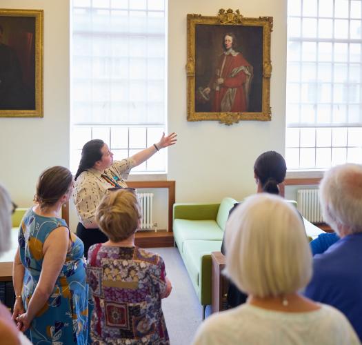 A group of visitors watch as a guide gestures towards a row of historic portraits in a room with large bright windows.