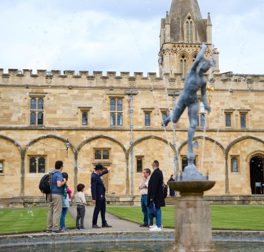 A group of visitors look around in a quadrangle surrounded by stone buildings, with a fountain in the centre.