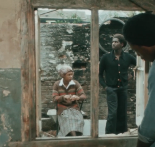 A woman sits and a man stands behind a wooden window frame.