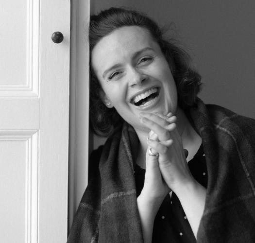 A black and white photograph of a woman laughing. Her hands are clasped together and she is leaning against a wooden panel containing a door.