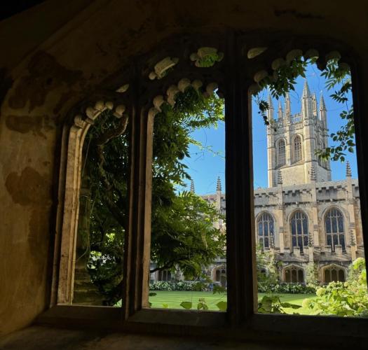 A medieval stone building with arched windows and a tower behind, viewed through a stone cloister looking out onto a lawn.