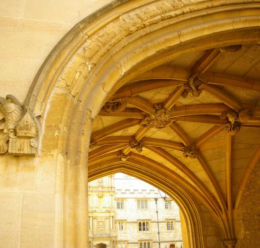 The archway of a medieval gatehouse with a vaulted ceiling and a stone college building beyond.