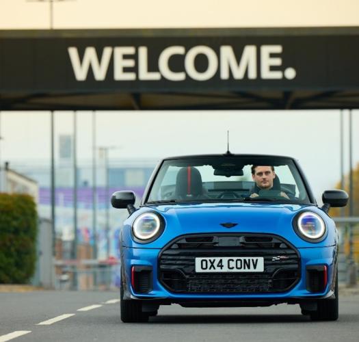 A blue Mini drives towards the camera beneath a black sign with Welcome written in white.