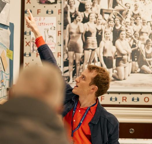 A guide in an orange shirt points at a history display during a tour to members of the public.
