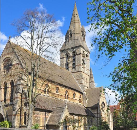 A sandy-coloured church with tower and steeple in a walled churchyard.