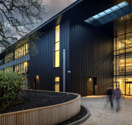 A black modern building with a large full height window and sweeping landscaping.