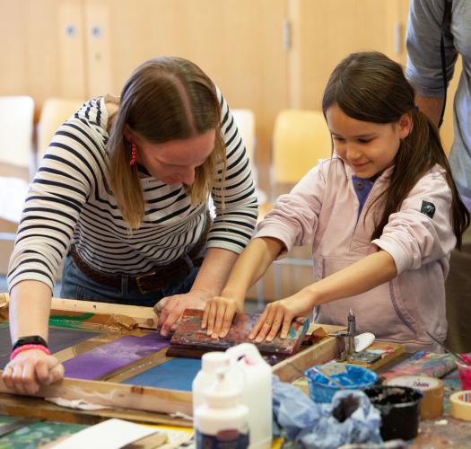 A woman in a stripey top held a girl scrape excess paint off a silk-screen printing frame. They are surrounded by paint pots and a boy looks over the girl's shoulder.