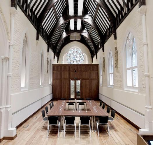 A chapel interior with bright white walls and dark wooden roof beams, with a conference style table in the centre.