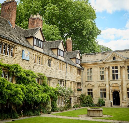 A sand-coloured stone building with gables and climbing foliage set around a grassy courtyard.