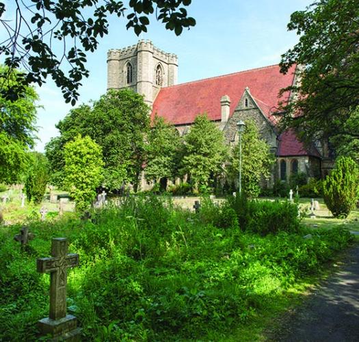 A lush churchyard with stone headstone and a path leading to a red roofed stone church with a tower.