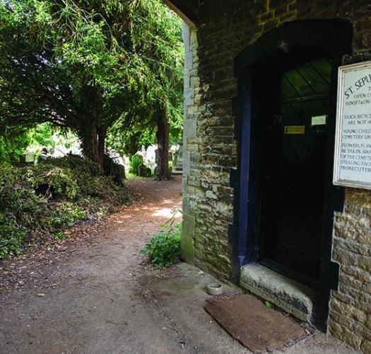 A path leading around a stone church building into a shaded tree-filled cemetery beyond.