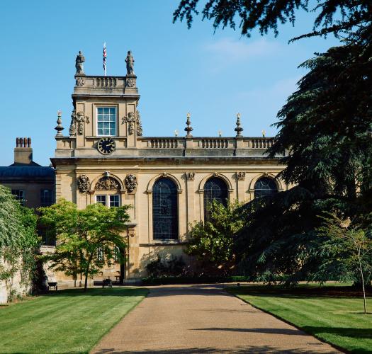 A wide path through a grassy area, leading to a seventeenth-century stone building with a tower, windows with rounded arches and an arched passageway through the building.