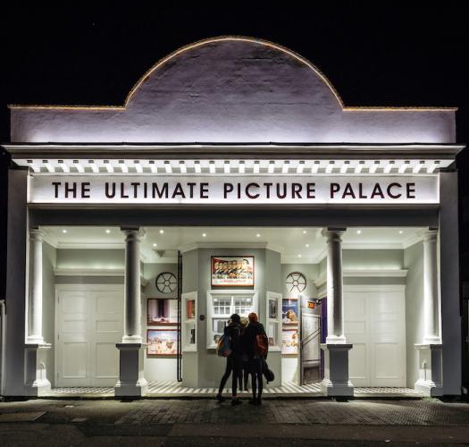 Two silhouetted figures stand in front of the illuminated facade of a white building with pillars and a sign reading The Ultimate Picture Palace. 