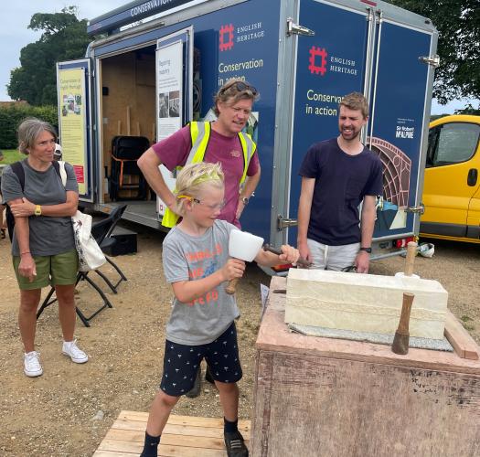 A boy has a go at stone carving with a chisel and mallet, with three adults standing watching next to an English Heritage Conservation in Action van.