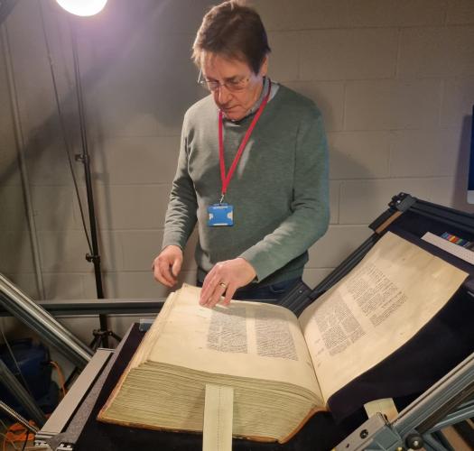 A man touches the edge of a page of a very large book, held in a cradle and lit by a very bright light.