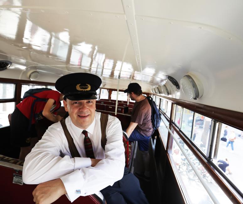 A man wearing a hat that says inspector looks at the camera inside a vintage bus with a white ceiling.