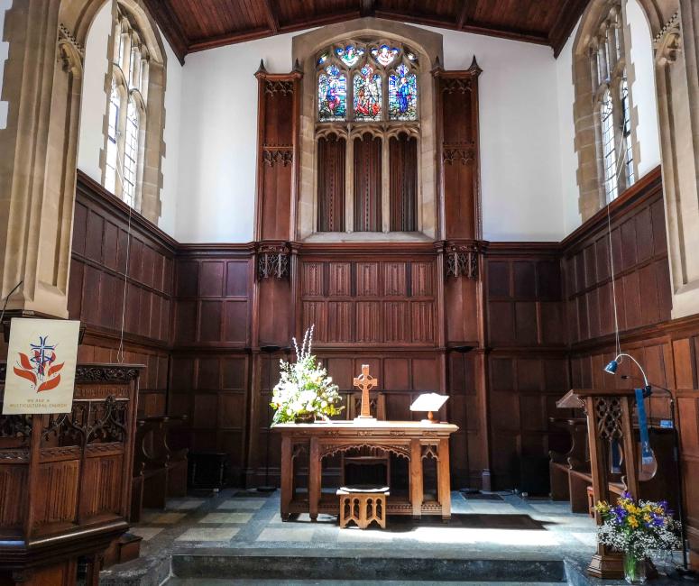 A wood-panelled church chancel with high windows on three sides and a wooden cross on a table in the centre.