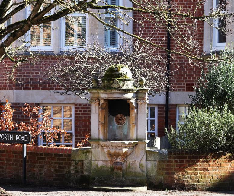 An ornate Victorian stone fountain set between two low brick walls, with trees overhead and brick houses in the background.