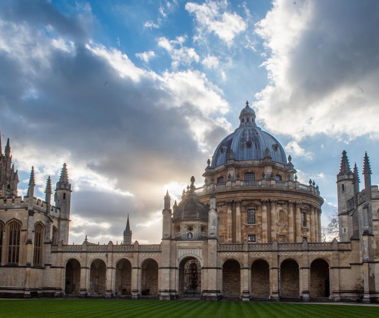 A stone university building with nine arches and a large round stone building behind. The sky is full of large clouds backlit by the sun.