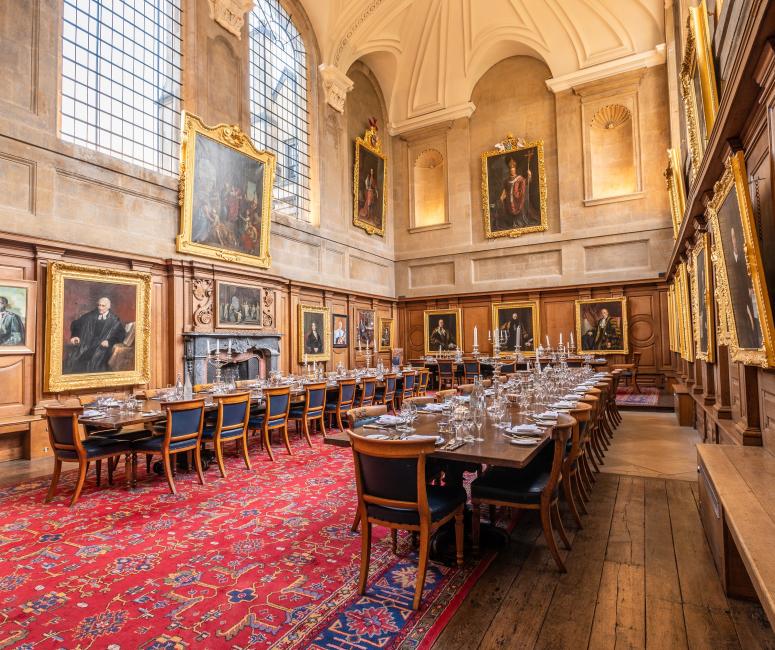 A large dining hall with long wooden tables, portraits hanging on stone walls and a long red carpet on the floor.