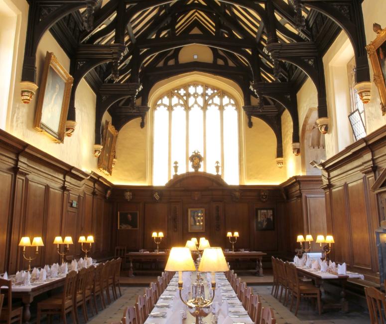 A tall dining hall with dark roof beams and a arched stone window at the end. There is a long table with a white table cloth and many place settings.