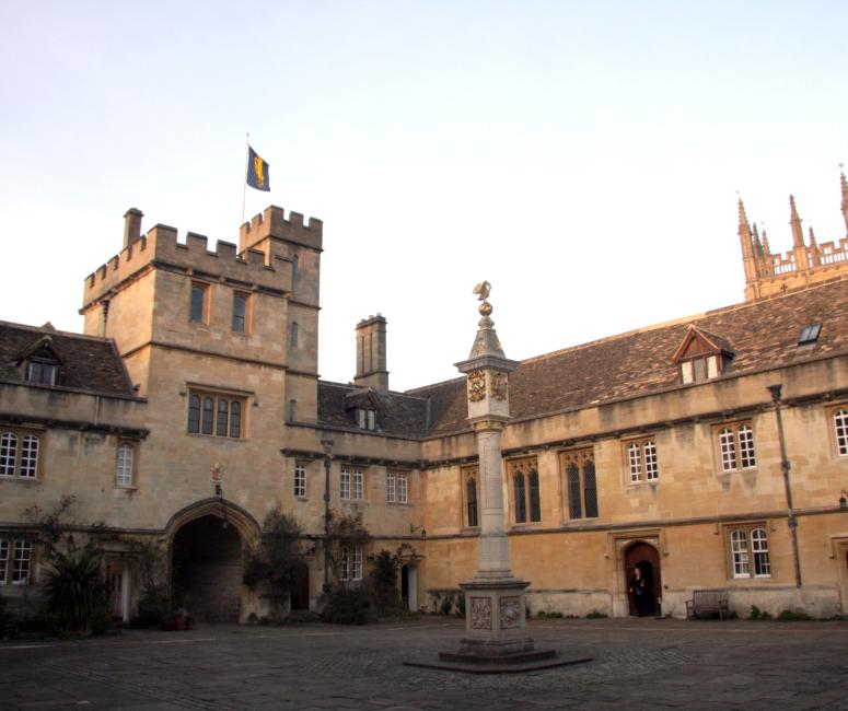 A sandy-coloured stone building with a gatehouse set around a paved courtyard with a column supporting a clock at the centre.