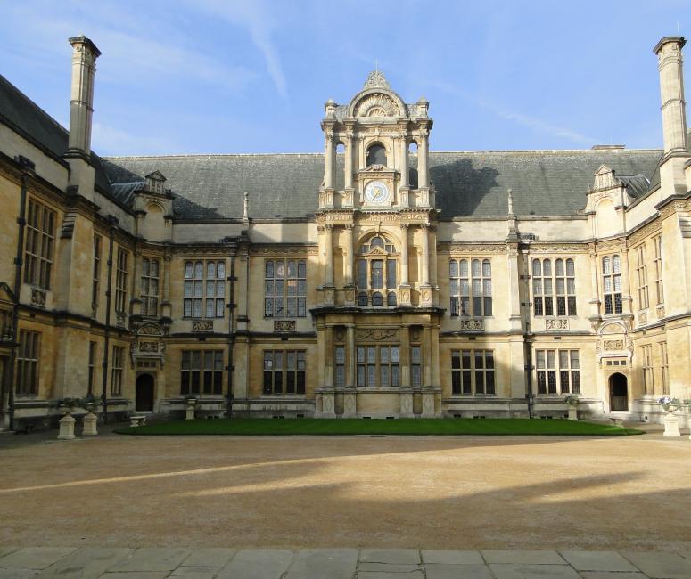 A stone building set around a gravel courtyard with a clock on the central part of the facade.