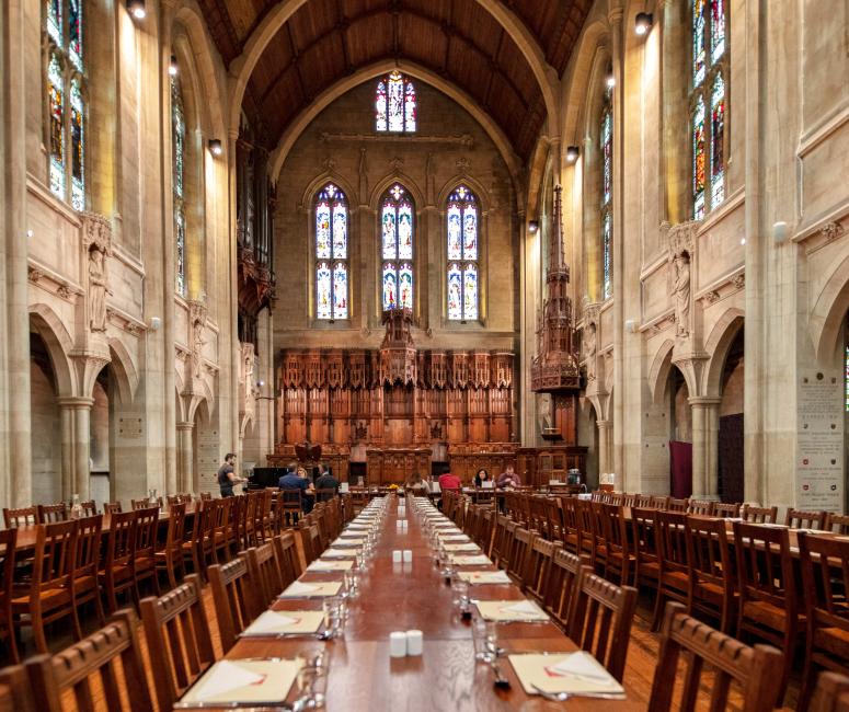 A large stone interior with a barrel vaulted roof and stained glass windows. Long wooden tables and chairs fill the room.